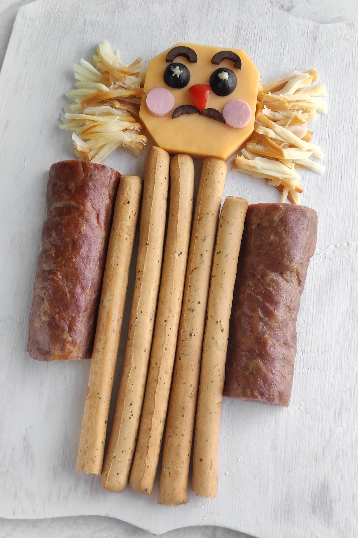 Nutcracker figure made from cheese slice face with olive eyes, shredded cheese hair, breadstick body, and meat roll arms.