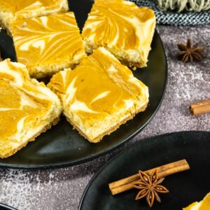 Pumpkin cheesecake bars with orange and cream swirled tops on a black plate.