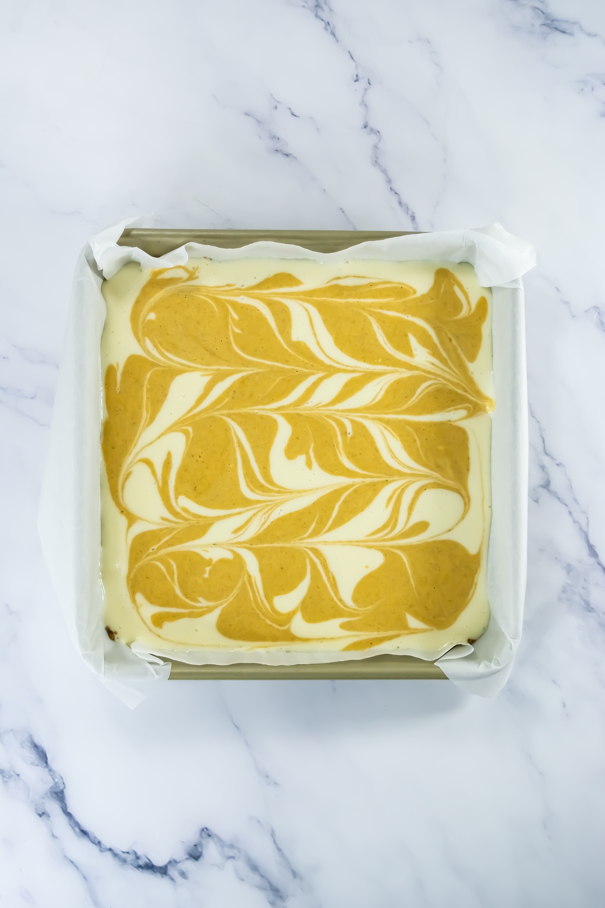 Unbaked pumpkin cheesecake batter in a parchment-lined square pan with orange pumpkin mixture swirled into cream cheese layer.