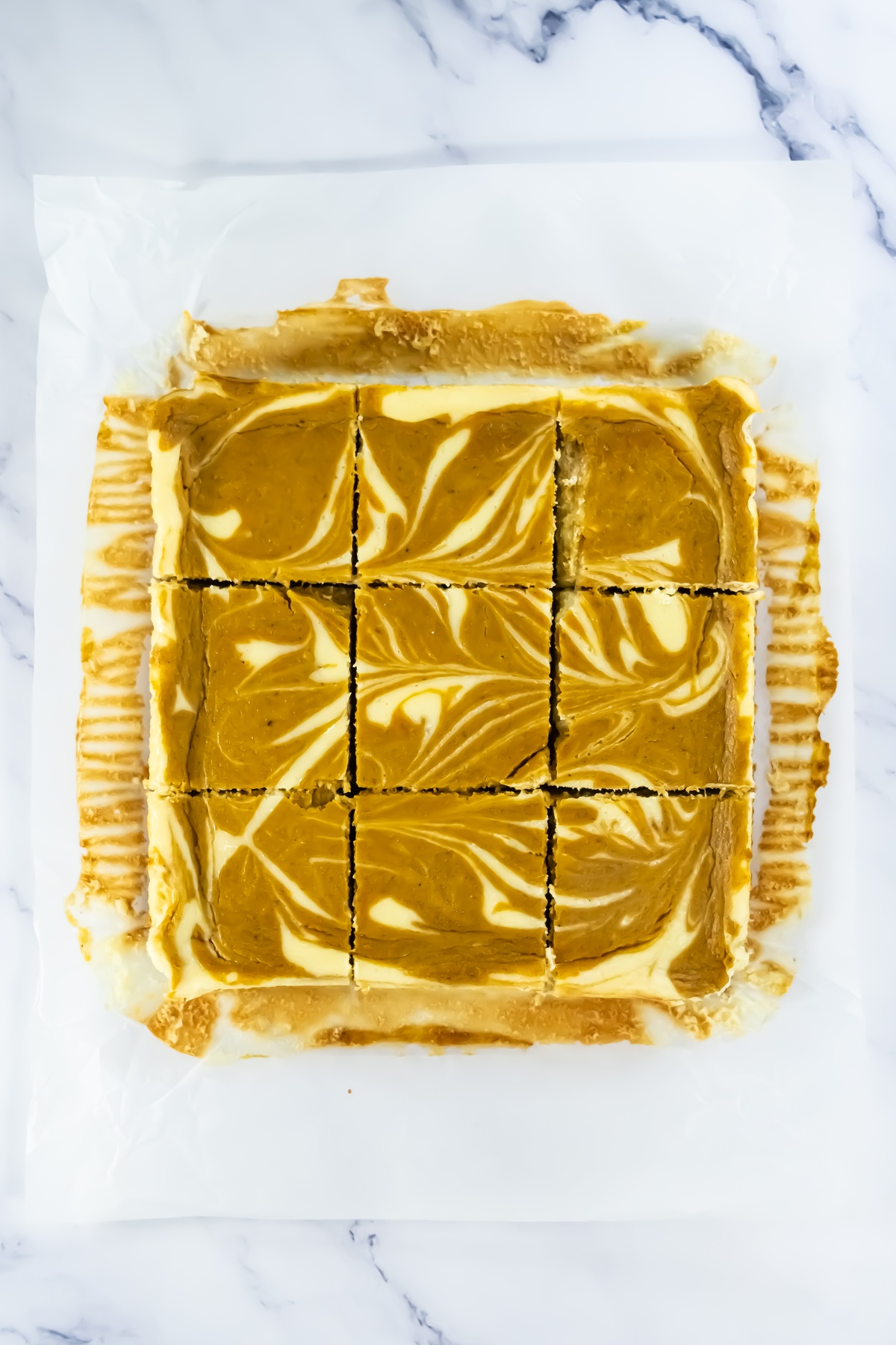 Baked pumpkin cheesecake bars with orange and cream swirl pattern, cut into 9 squares on parchment paper.