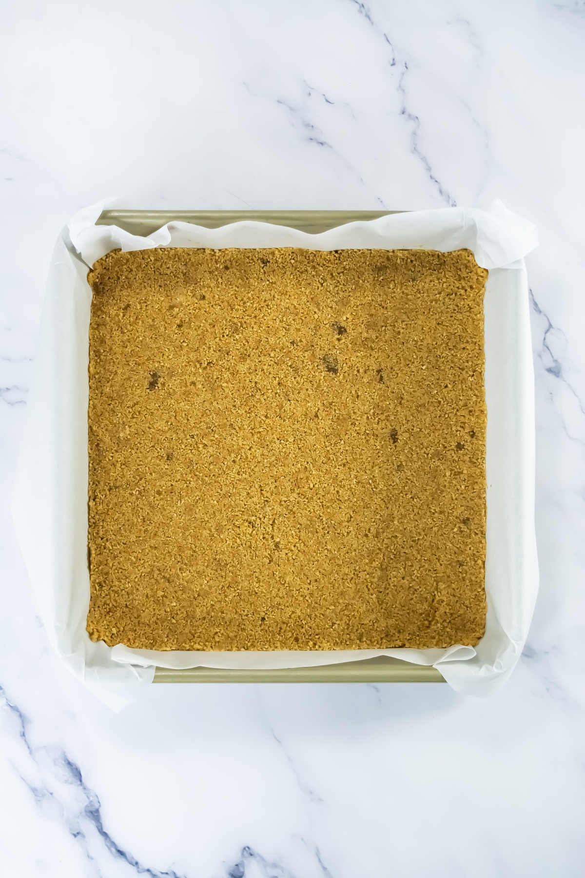 Golden graham cracker crust pressed evenly into a parchment-lined square baking pan on a marble surface.