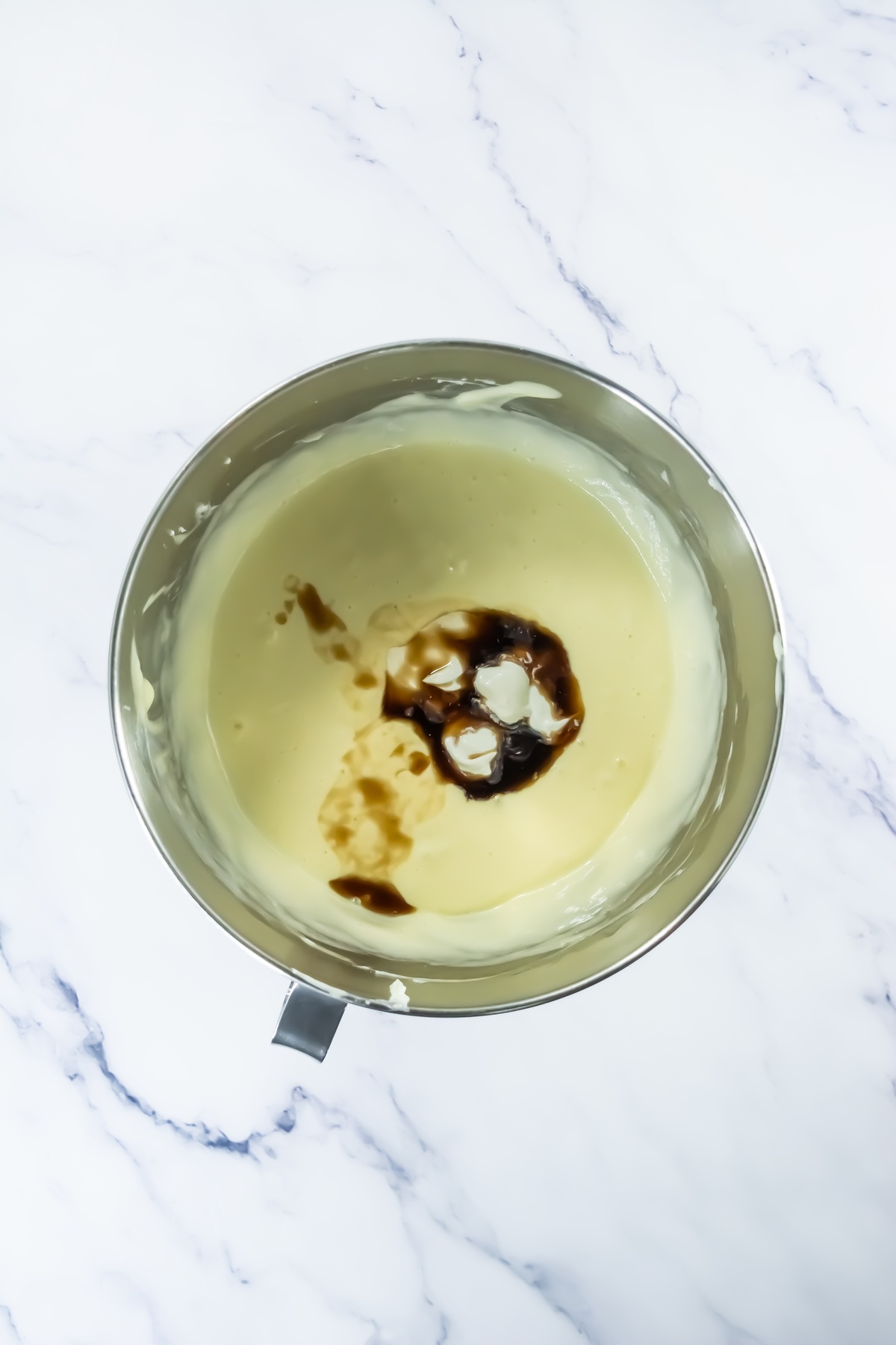 Stainless steel mixing bowl containing pale creamy batter with vanilla extract added, ready to be mixed, on marble surface.