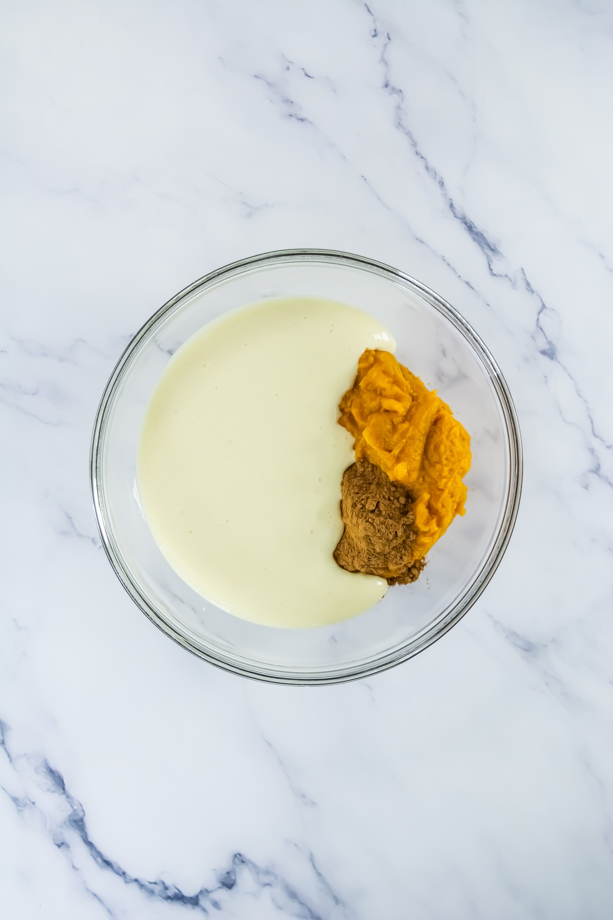 Glass mixing bowl with sweetened condensed milk, pumpkin puree, and spices before mixing on marble surface.