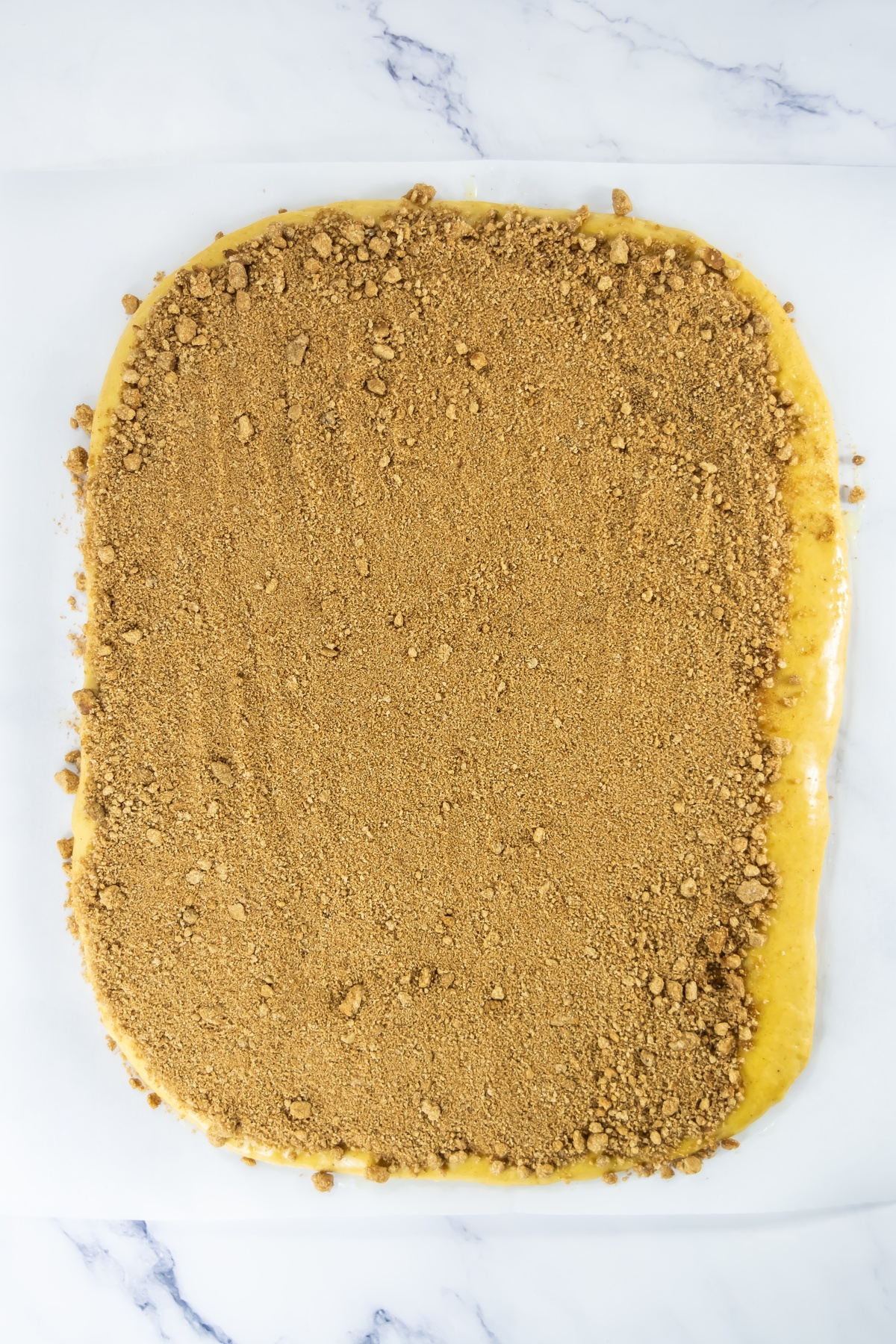 Rolled out yellow dough covered with brown sugar and cinnamon mixture on a marble surface, ready to be rolled.