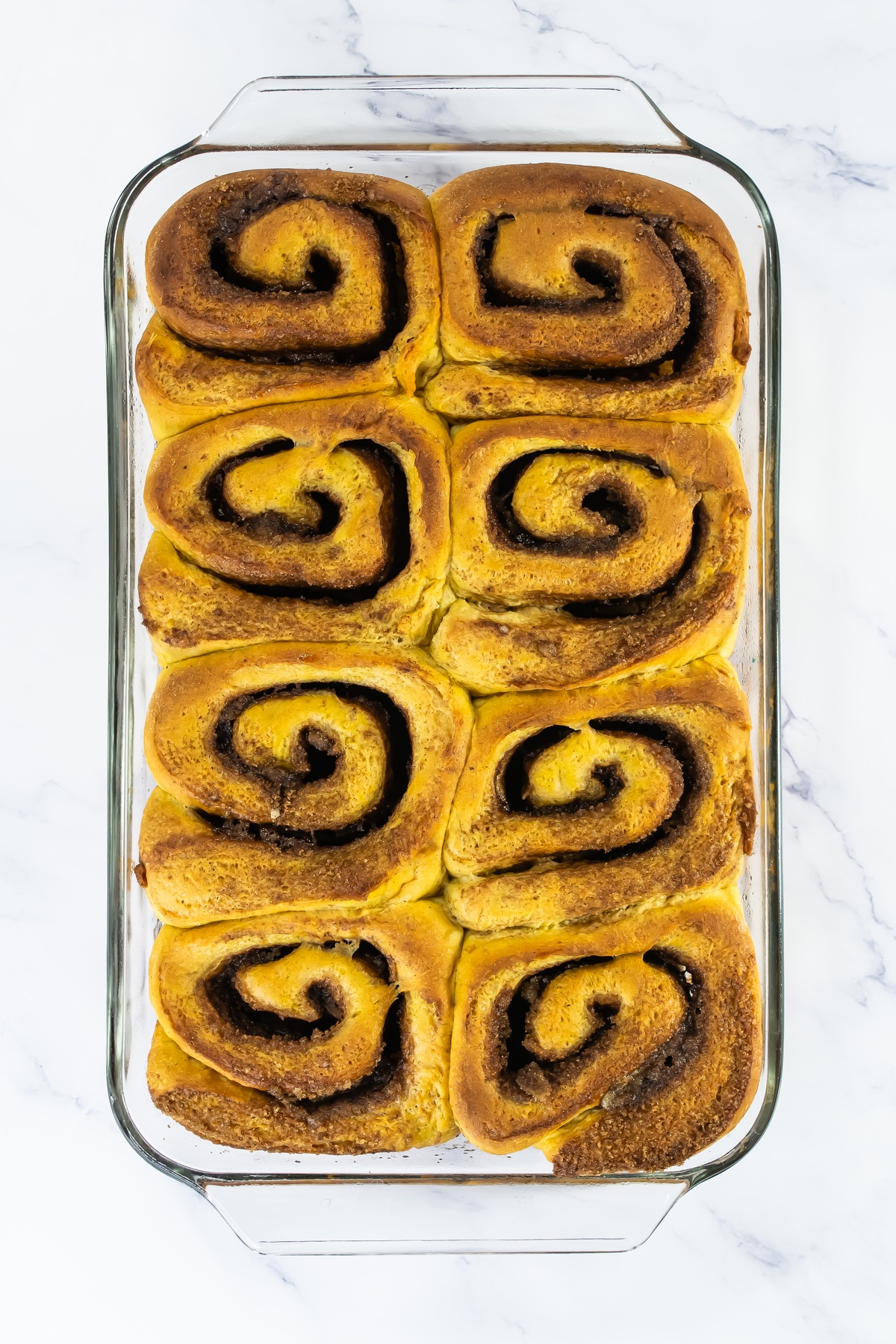 Freshly baked pumpkin cinnamon rolls with golden swirled tops arranged in a glass baking dish on a marble surface.