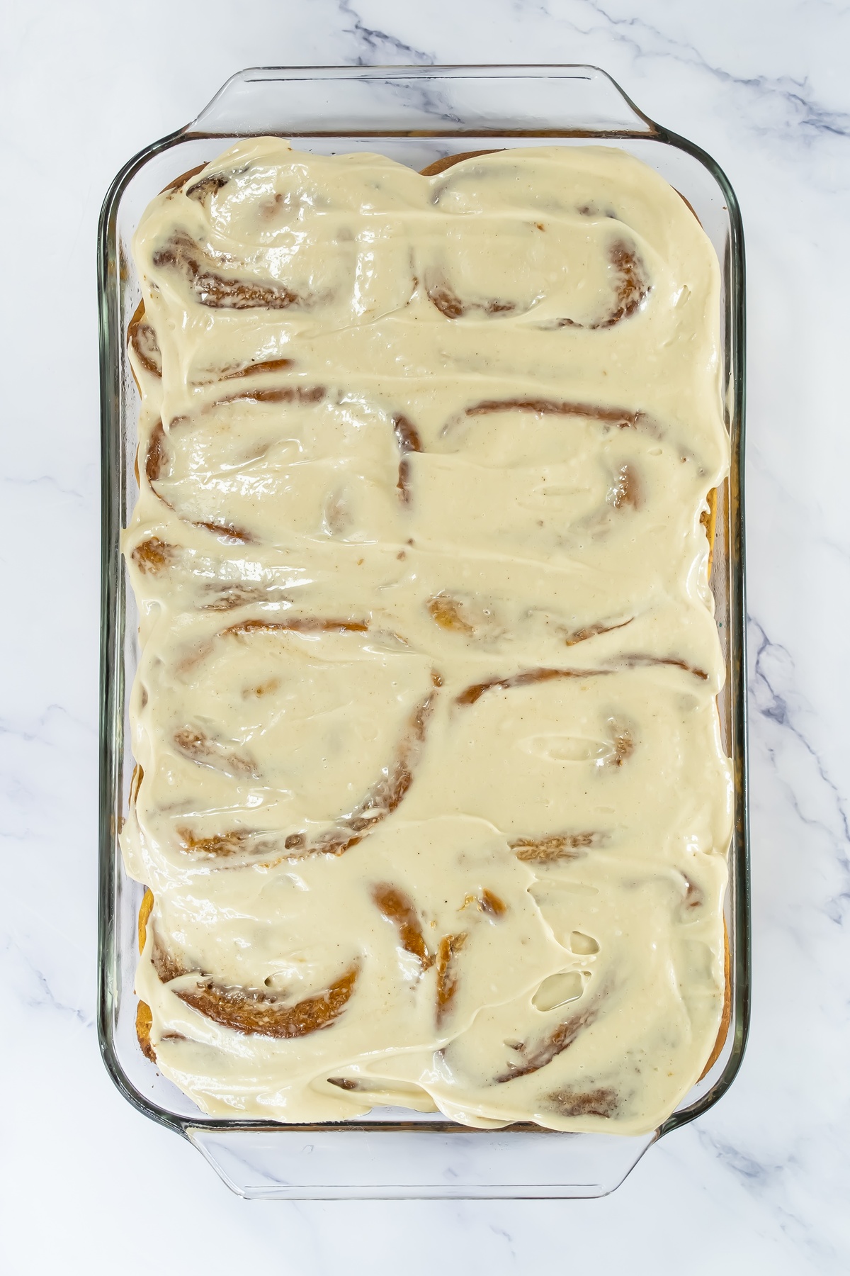 Freshly baked pumpkin cinnamon rolls topped with cream cheese frosting in a glass baking dish on a marble surface.