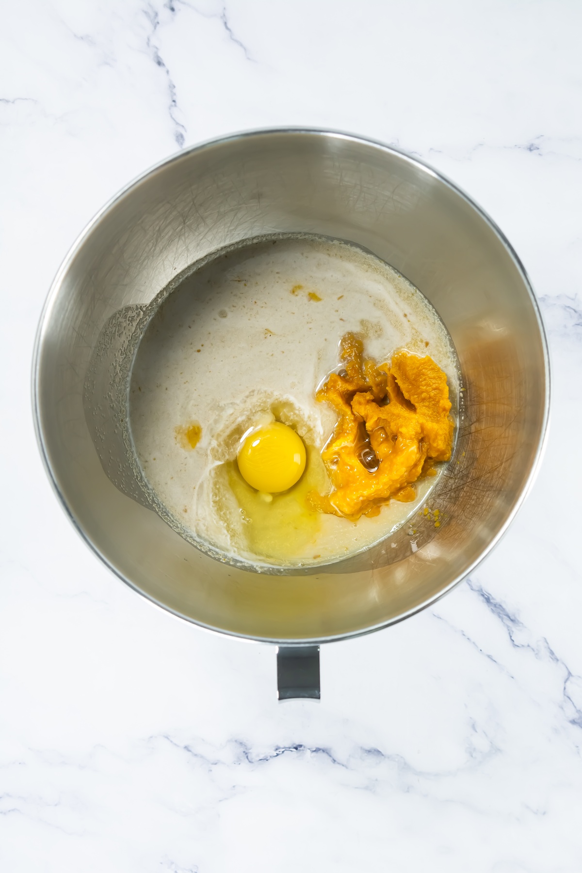 Stainless steel mixing bowl containing raw egg, pumpkin puree, and foamy yeast mixture on marble surface.