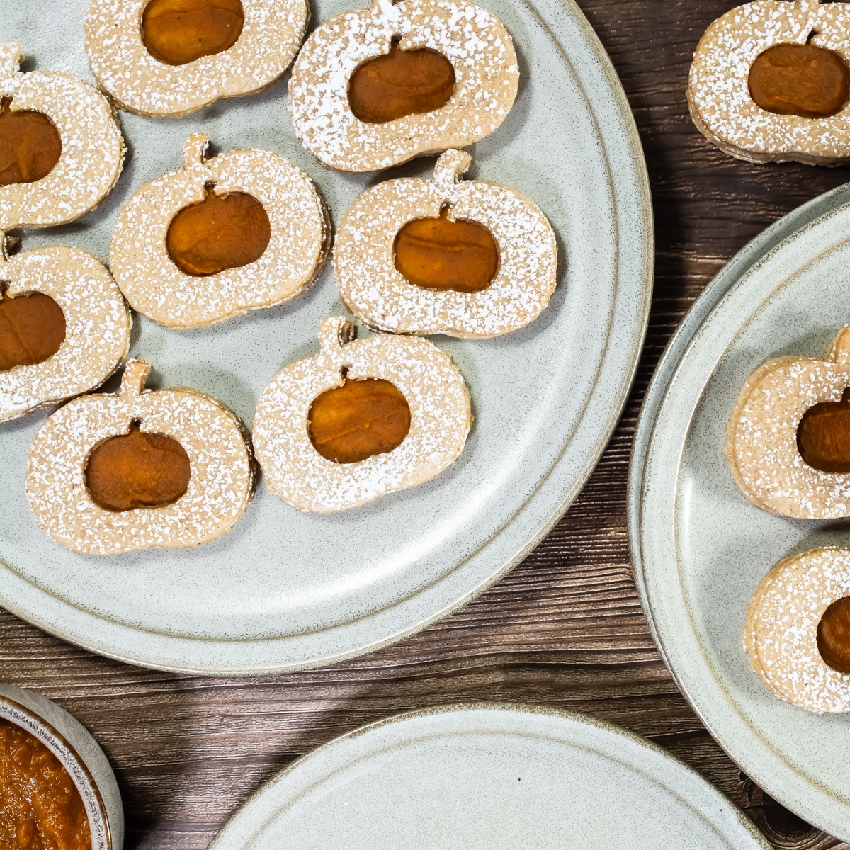 Pumpkin-shaped linzer cookies with orange filling and powdered sugar on light gray ceramic plates on a dark wood surface.