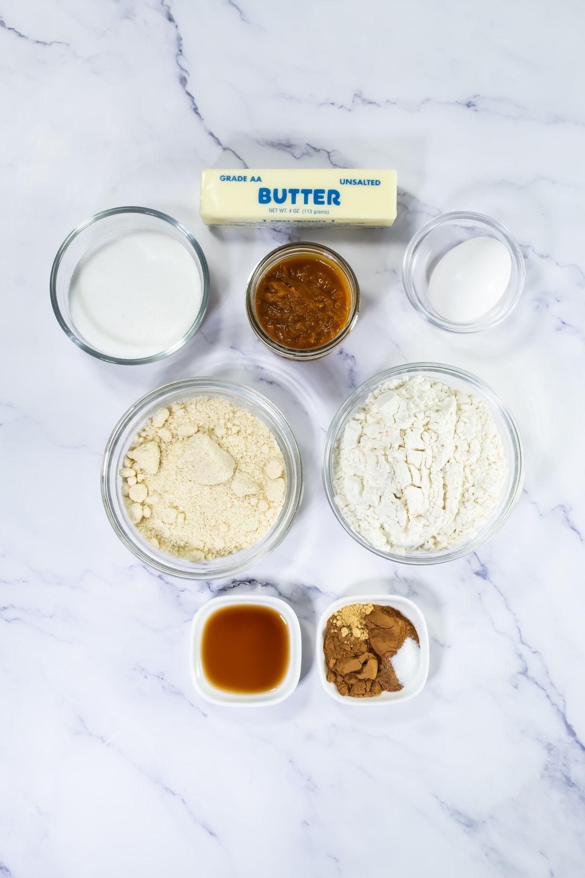 Baking ingredients on marble surface: stick of unsalted butter, bowls of milk, jam, egg, almond flour, white flour, vanilla.
