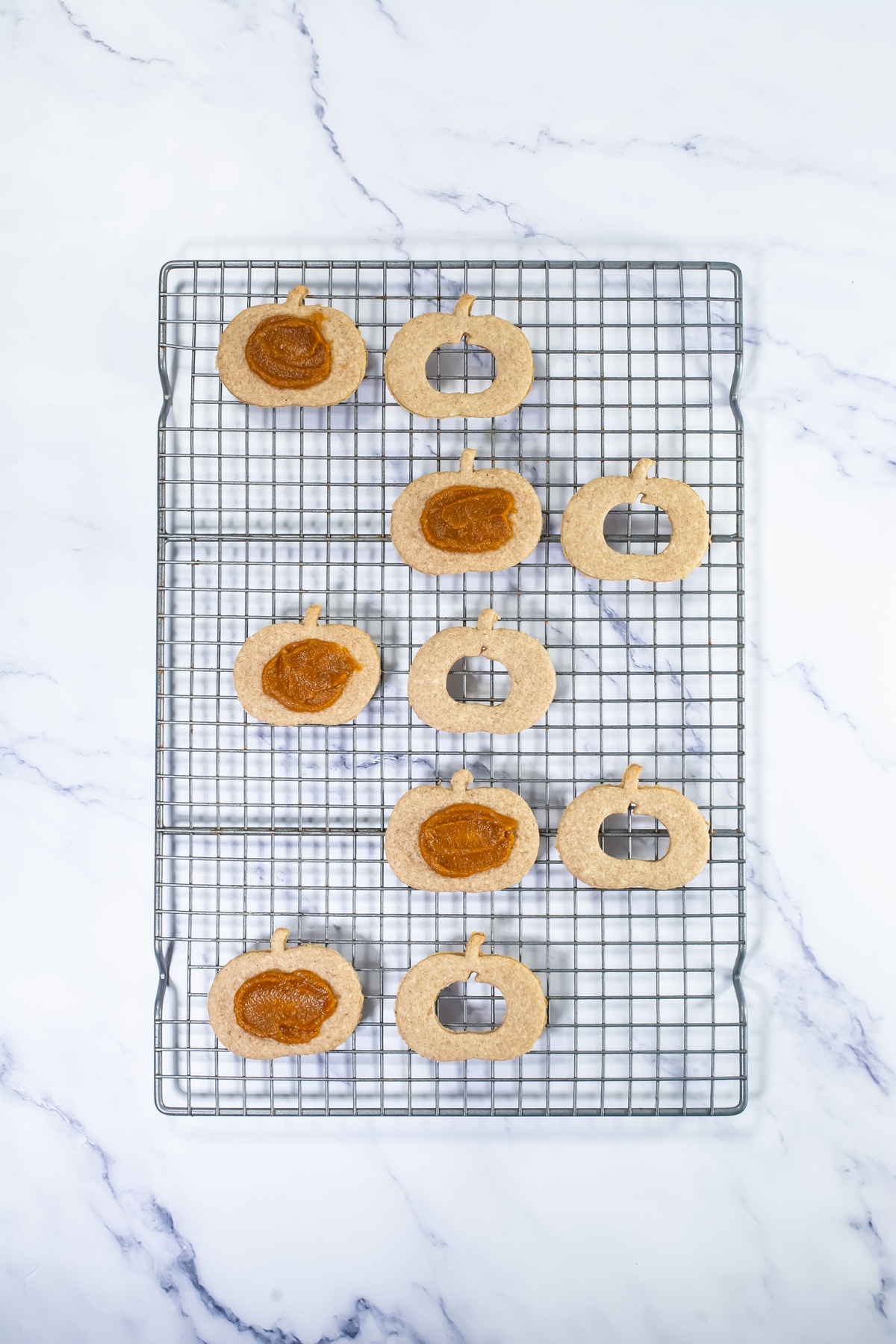 Baked pumpkin-shaped linzer cookies cooling on a wire rack, with solid bases topped with pumpkin filling.