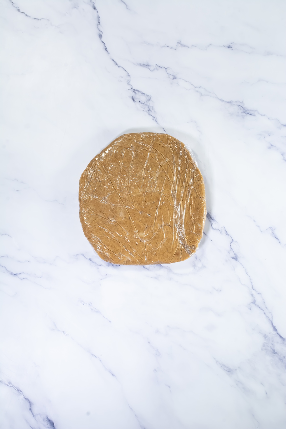 Flattened cookie dough wrapped in plastic wrap resting on a white marble surface, ready for chilling.