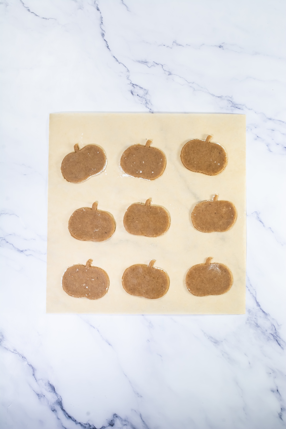 Nine unbaked pumpkin-shaped cookies arranged in rows on parchment paper on a marble surface.