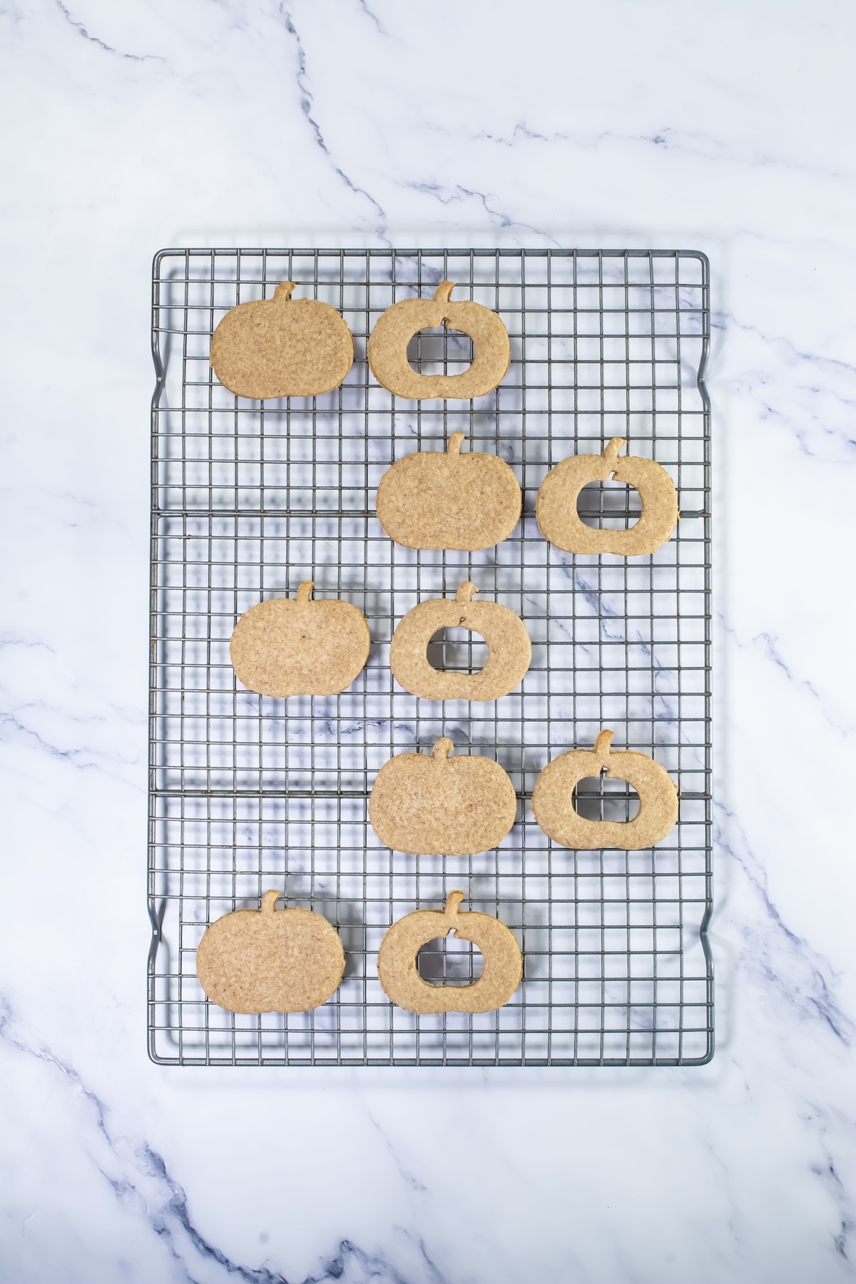 Ten baked pumpkin-shaped linzer cookies cooling on a wire rack, with half showing center cutouts, on a marble surface.