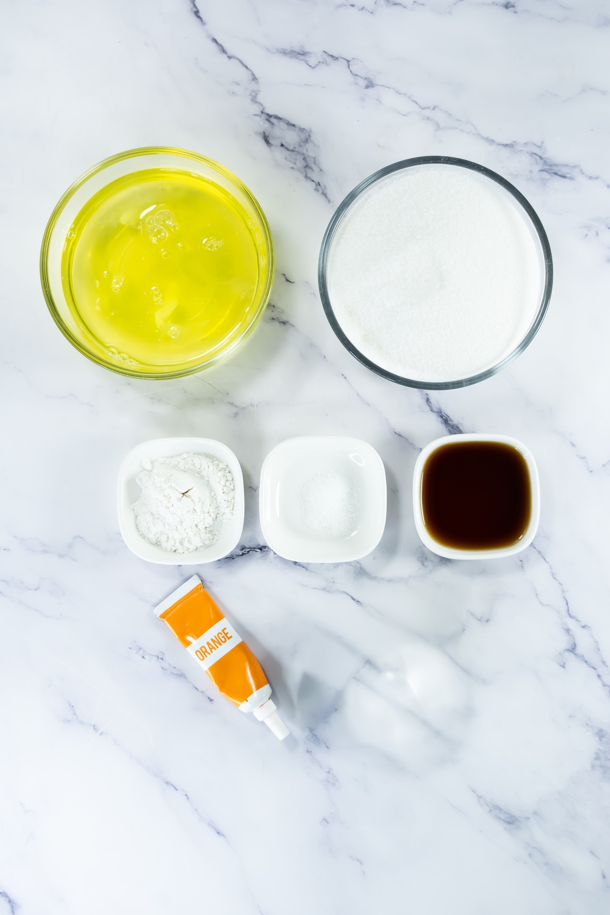 Ingredients for meringues: egg whites in glass bowl, sugar, cornstarch, salt, vanilla extract, and orange food coloring.