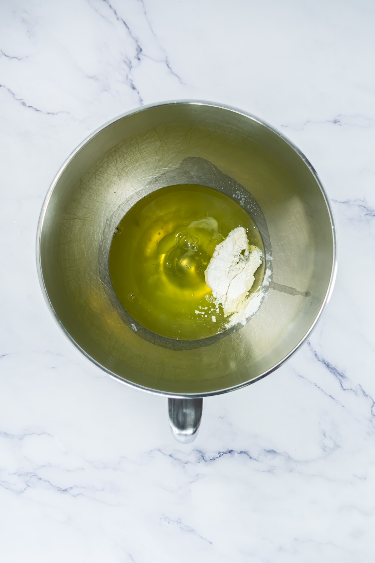 Egg whites and a white powdery ingredient in a stainless steel mixing bowl on a marble surface, unmixed.