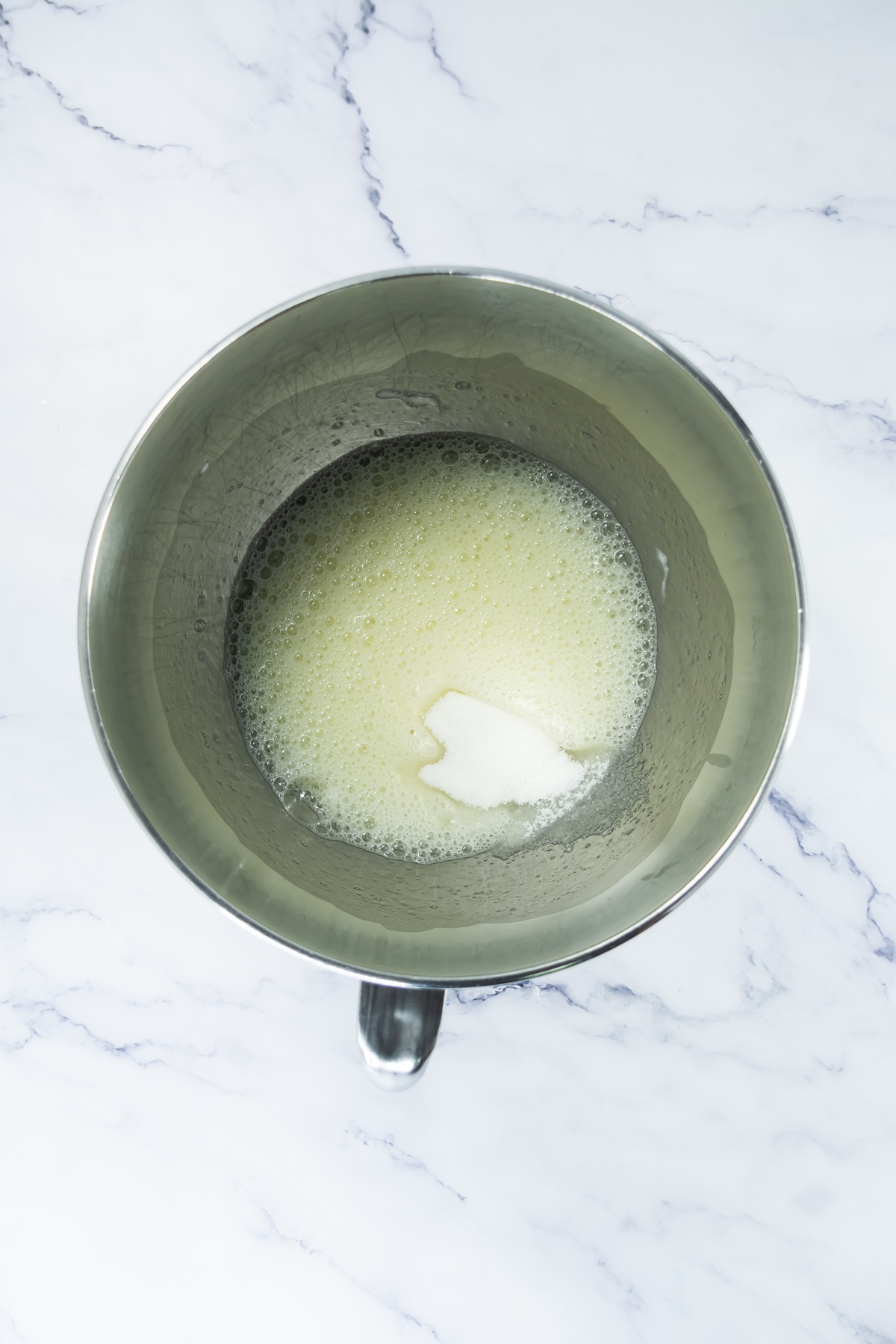 Foamy egg whites with sugar in a stainless steel mixing bowl on a marble surface.