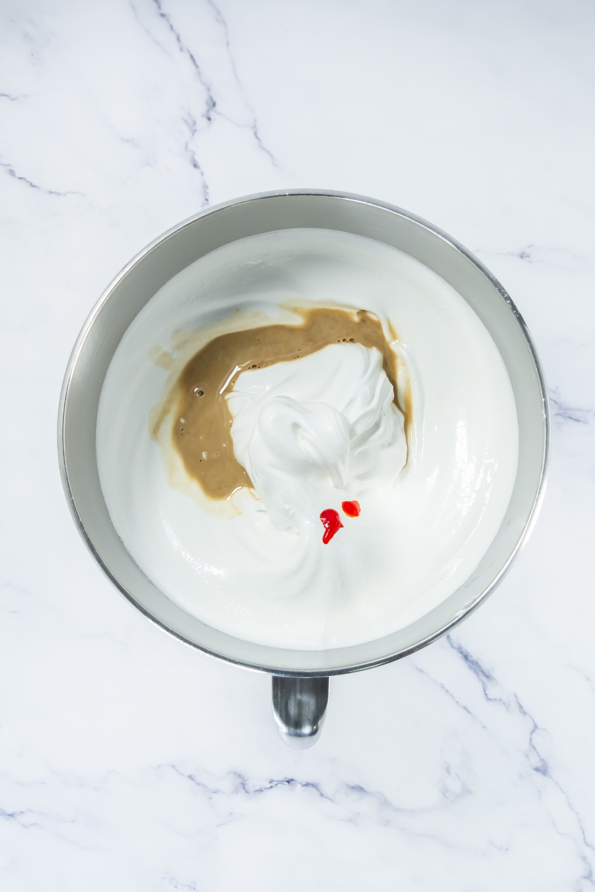 Stiff white meringue in a metal stand mixer bowl with brown liquid and drops of red food coloring added before mixing.