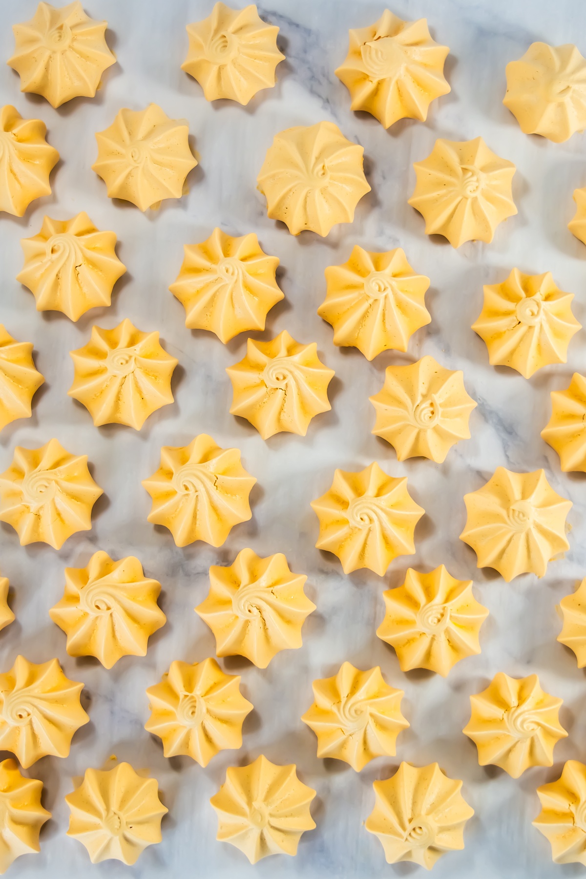 Rows of baked orange-yellow swirled meringue cookies with pointed peaks arranged on a marble surface.