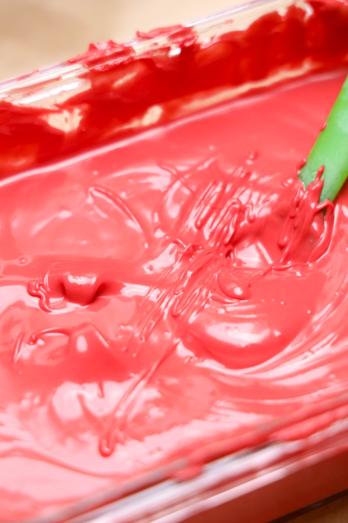 Pink melted candy coating in a clear glass dish with a green spatula, drizzled with darker pink lines on the surface.