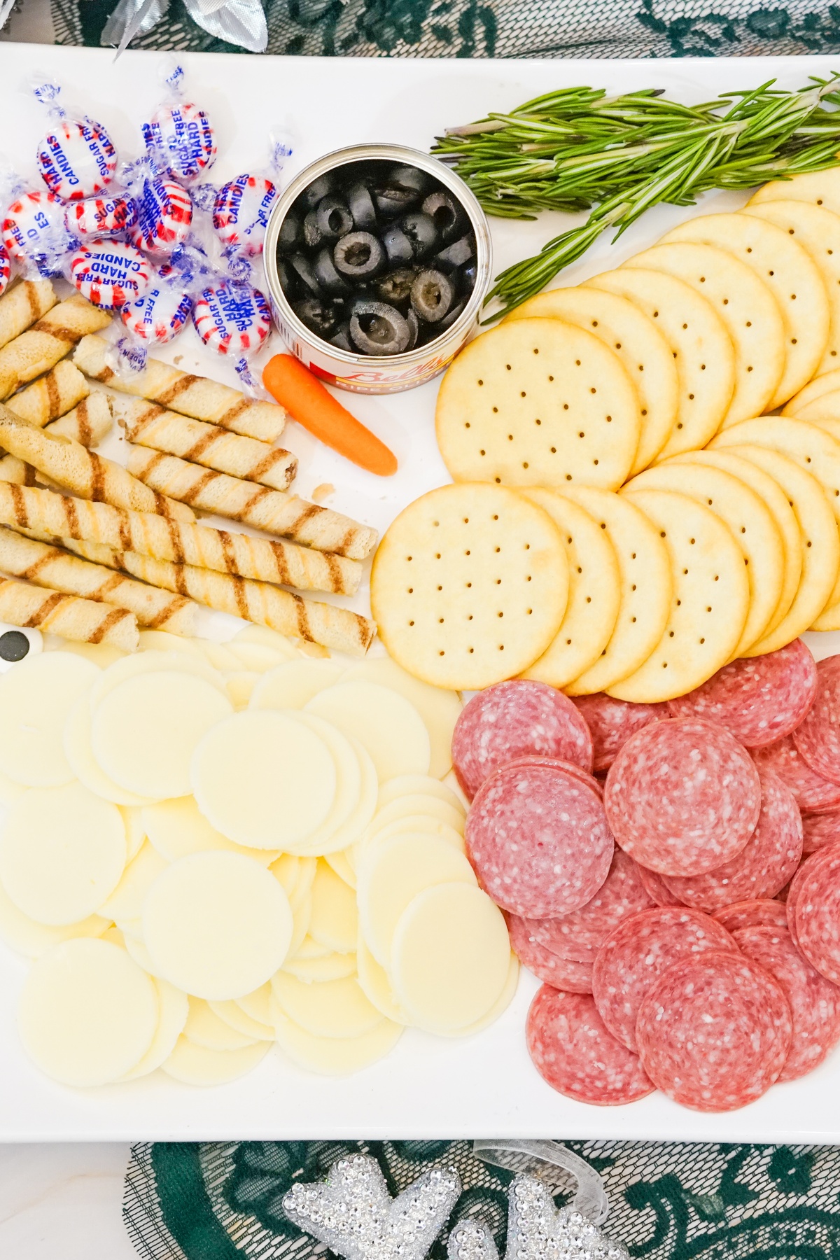 Charcuterie board ingredients laid out: sliced salami, round white cheese, crackers, breadsticks, black olives in can.