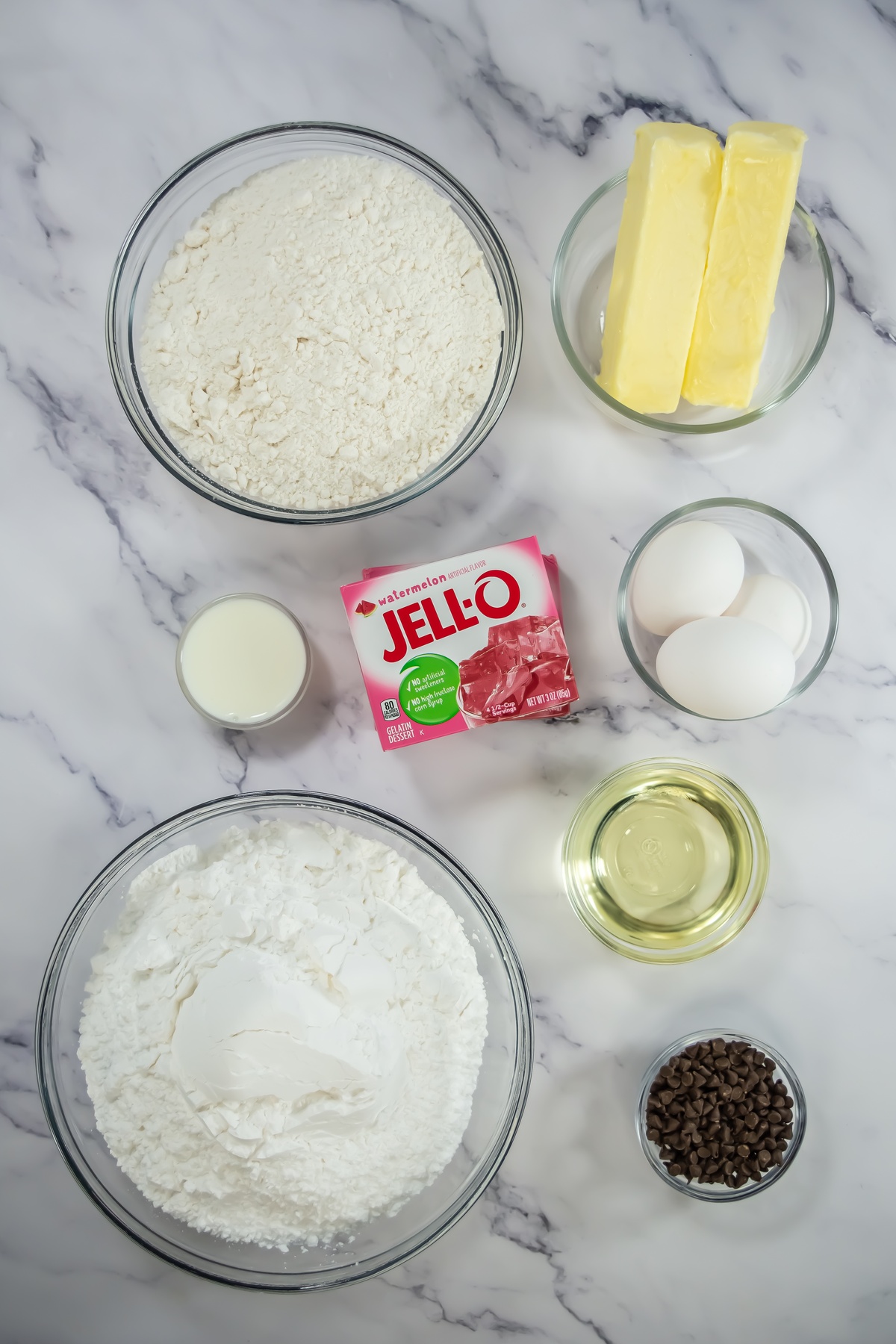 Baking ingredients on marble surface: flour, butter sticks, eggs, milk, watermelon Jell-O, powdered sugar, oil, and chocolate chips.