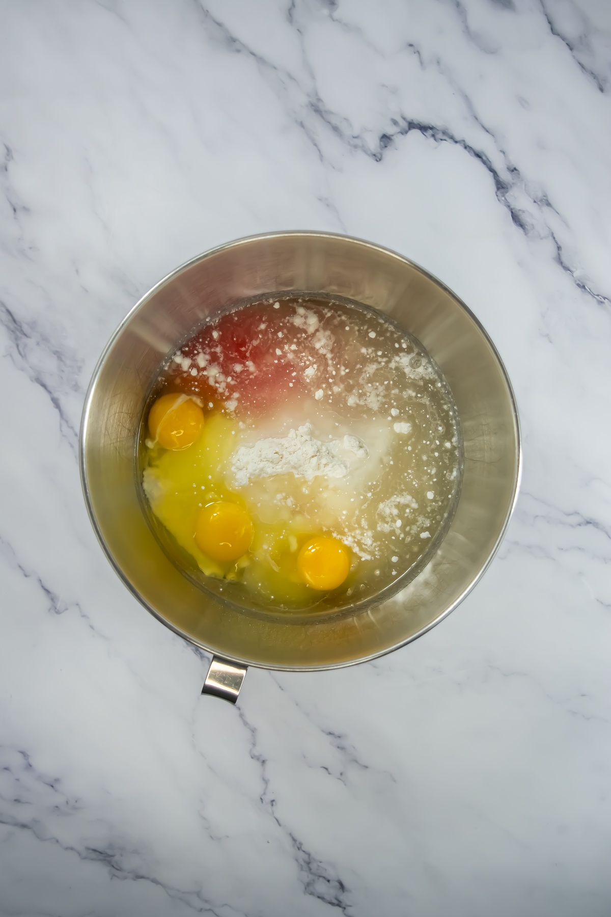 Stainless steel mixing bowl containing raw cracked eggs, flour, and liquid ingredients on white marble countertop.