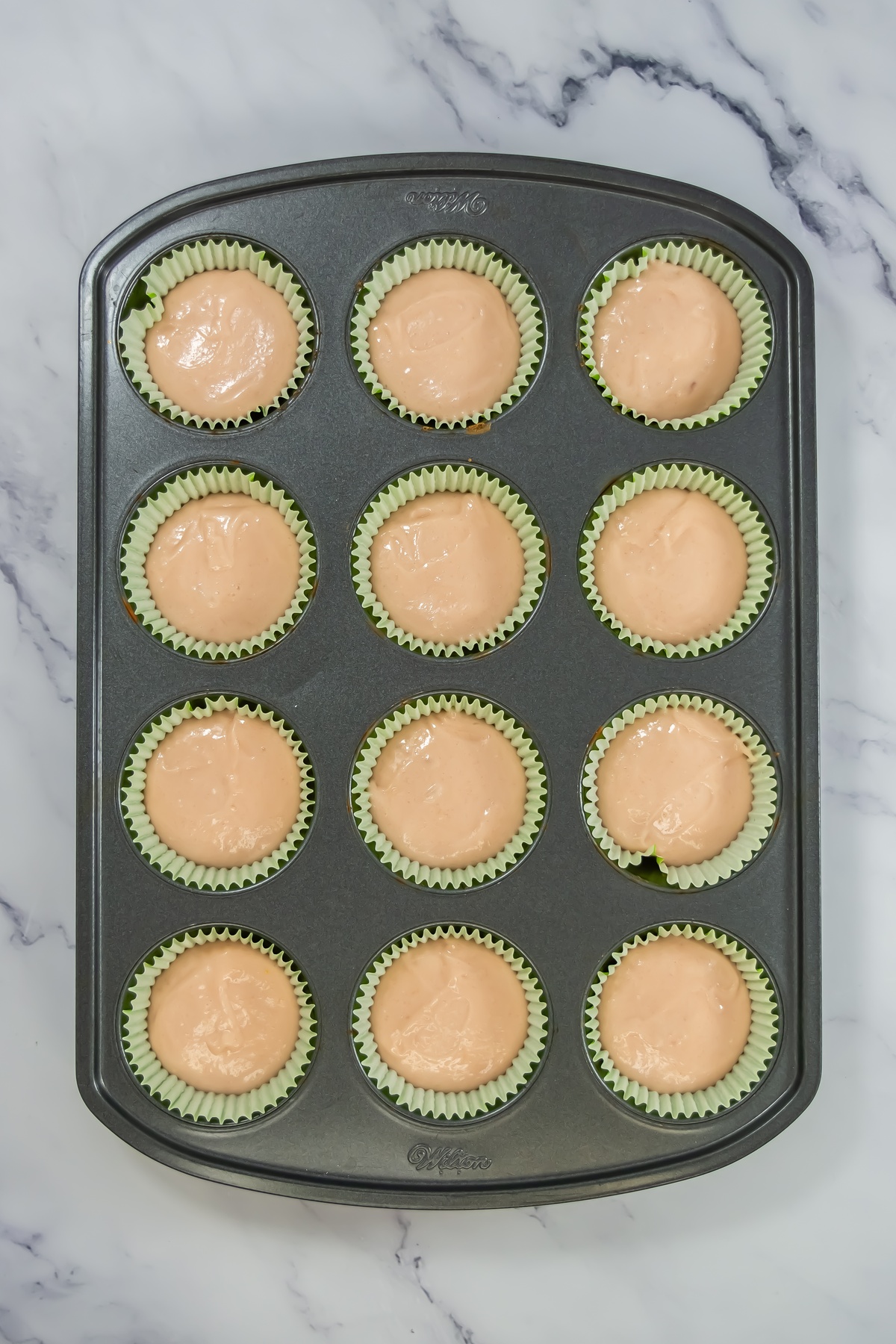Twelve cupcake liners filled with pink batter in a Wilton muffin tin, ready to bake, on a marble surface.
