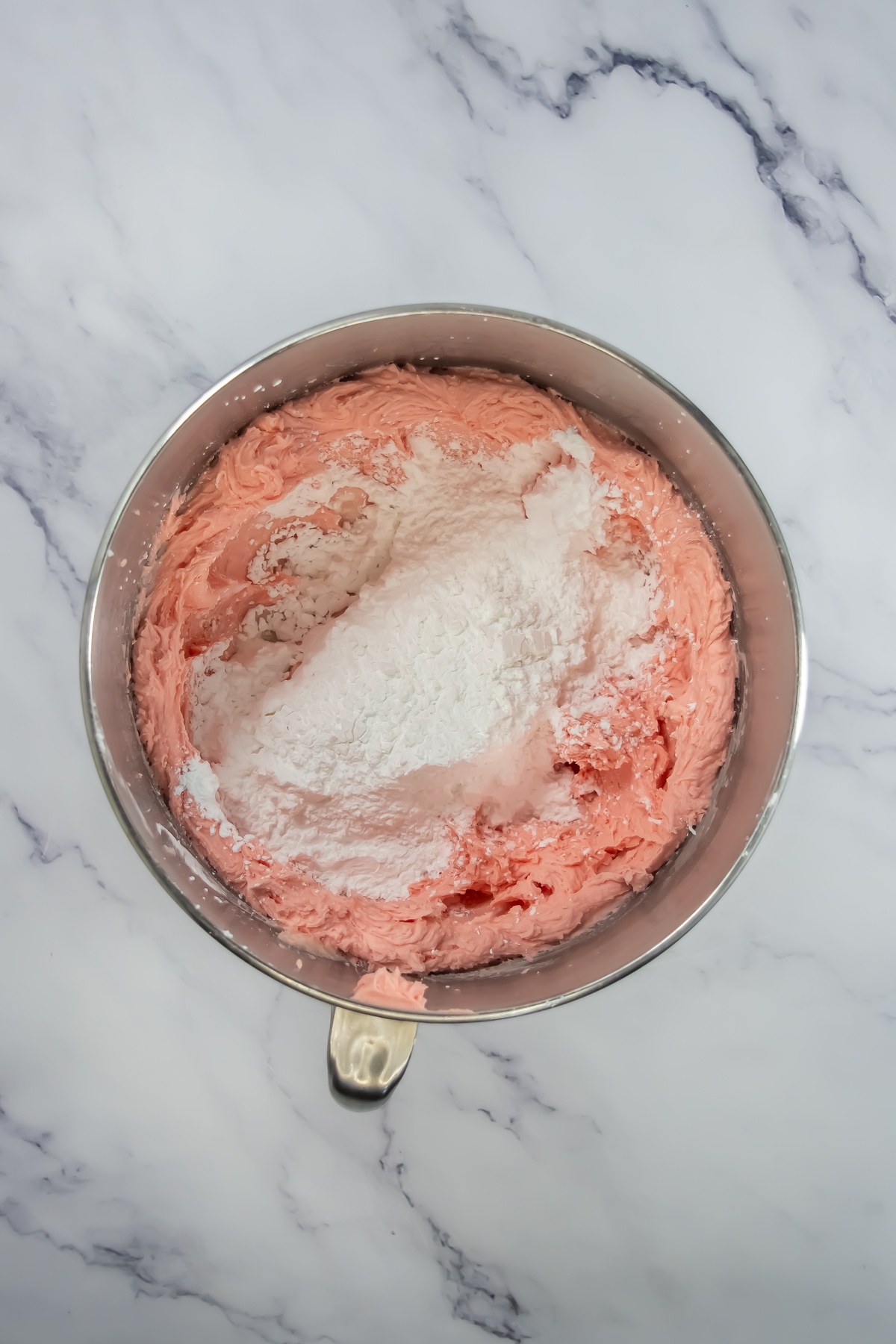 Pink frosting in a metal stand mixer bowl with powdered sugar added on top, ready to be mixed.