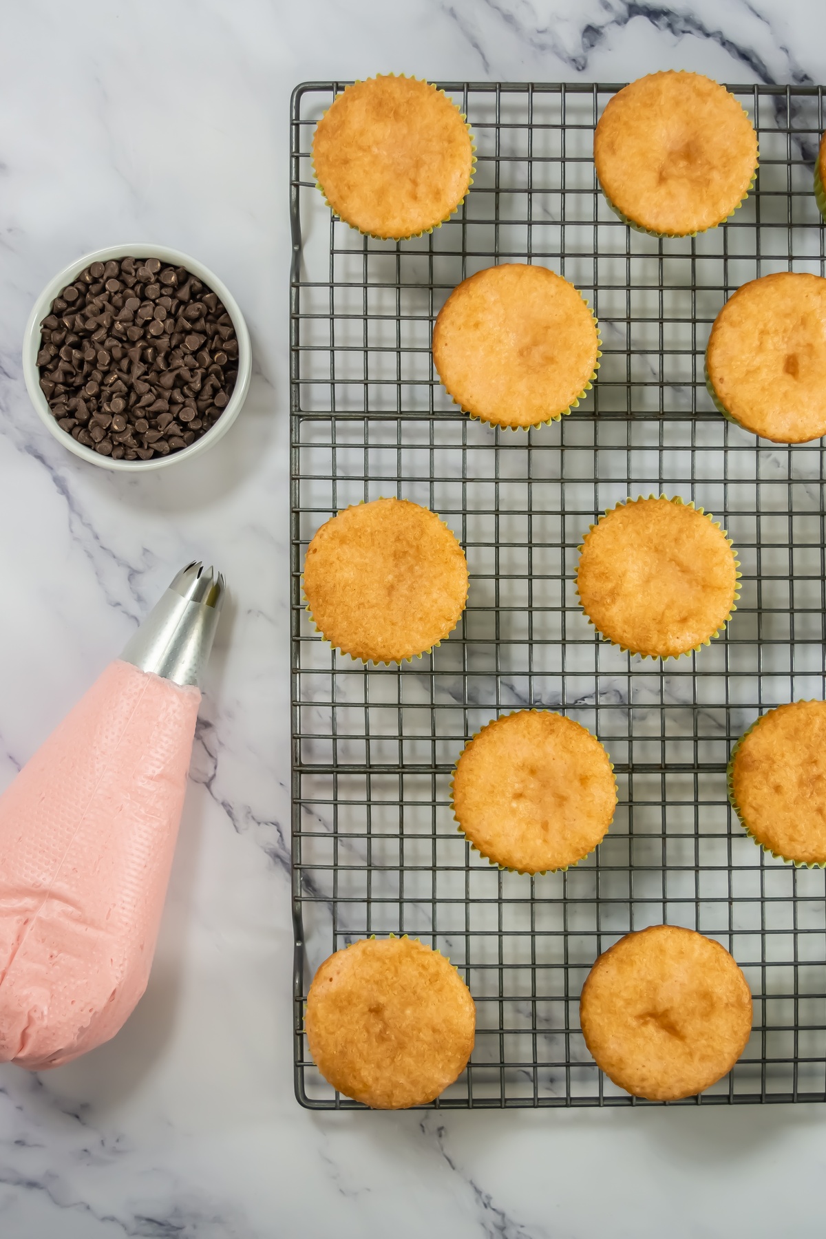 Baked vanilla cupcakes cooling on a wire rack beside a bowl of mini chocolate chips and a piping bag filled with pink frosting.